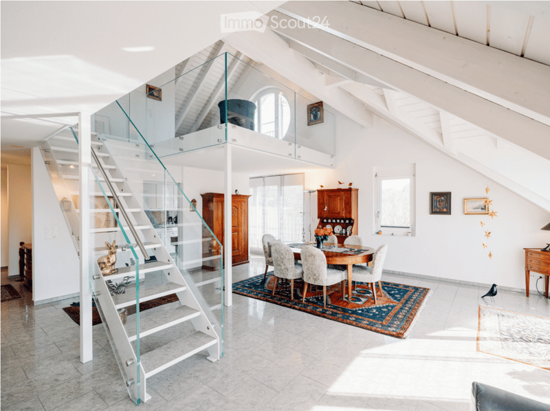 Modern, open-concept living and dining area with high ceilings, exposed wooden beams, and a glass staircase leading to an upper level. The dining area features a round wooden table with six upholstered chairs, a colorful area rug, and a vase with flowers