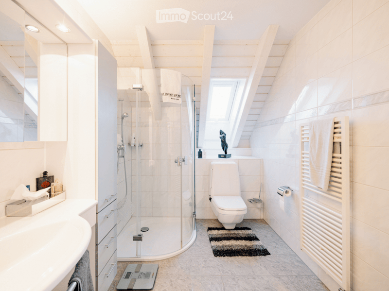 Modern bathroom with white tiles, a glass-enclosed shower, a toilet, a sink with a mirror, a towel rack with a towel, a decorative sculpture on the windowsill, and a scale on the floor.