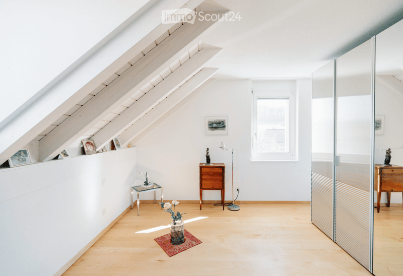 Bright attic room with wooden floors, large skylights, white walls, small table, chair, vase with flowers, picture frames, and floor lamp.
