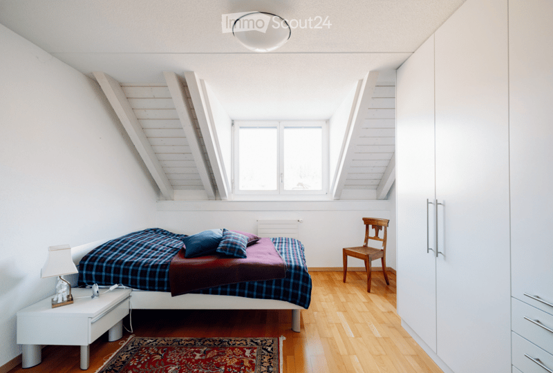Bright, modern bedroom with wooden flooring, white walls, and a slanted ceiling. Features a neatly made bed with plaid bedding, a nightstand with a lamp, a wooden chair, and a built-in wardrobe.