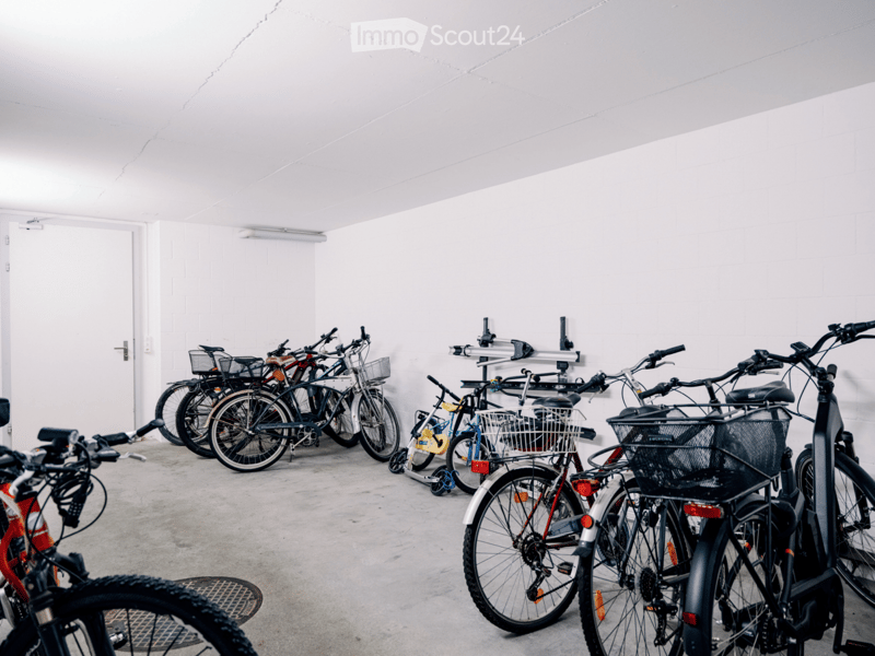Empty indoor parking area with several bicycles stored in racks against a white wall. The space has a concrete floor and a white ceiling with a single light fixture.