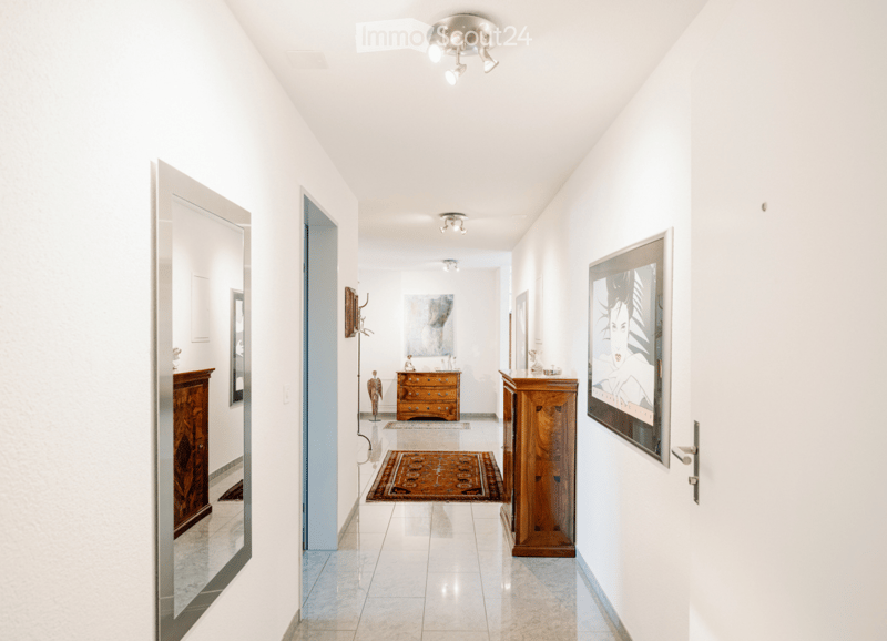 Bright, modern hallway with white walls, tiled floor, recessed lighting, and several doors leading to rooms. Features include a large mirror, artwork, and small wooden cabinets.