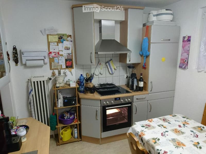 Functional kitchen with modern appliances, including an oven, stove, refrigerator, and cabinets. A small table with a floral tablecloth and a shelf with various items are also visible.