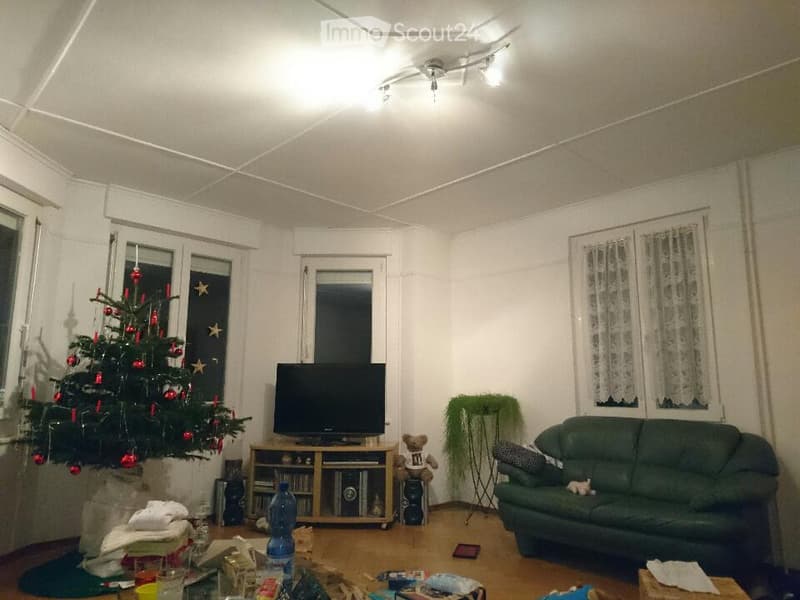 A cozy living room with a green sofa, a television on a wooden stand, a Christmas tree, and various items scattered around, including a bottle and cups on a coffee table.