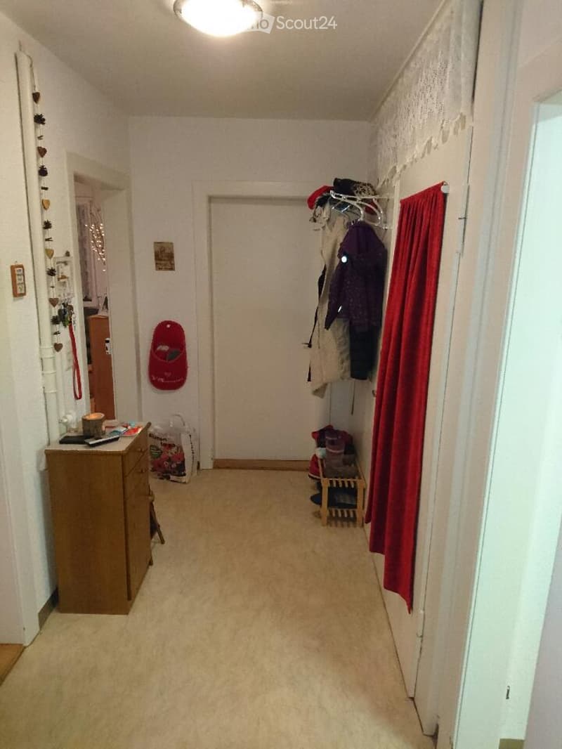 A narrow hallway with beige carpeting and white walls. A wooden dresser with a cup and remote control sits on the left, while a coat rack with jackets and a red bag hangs on the right. A small wooden stool with items on it is near the coat rack, and a pic