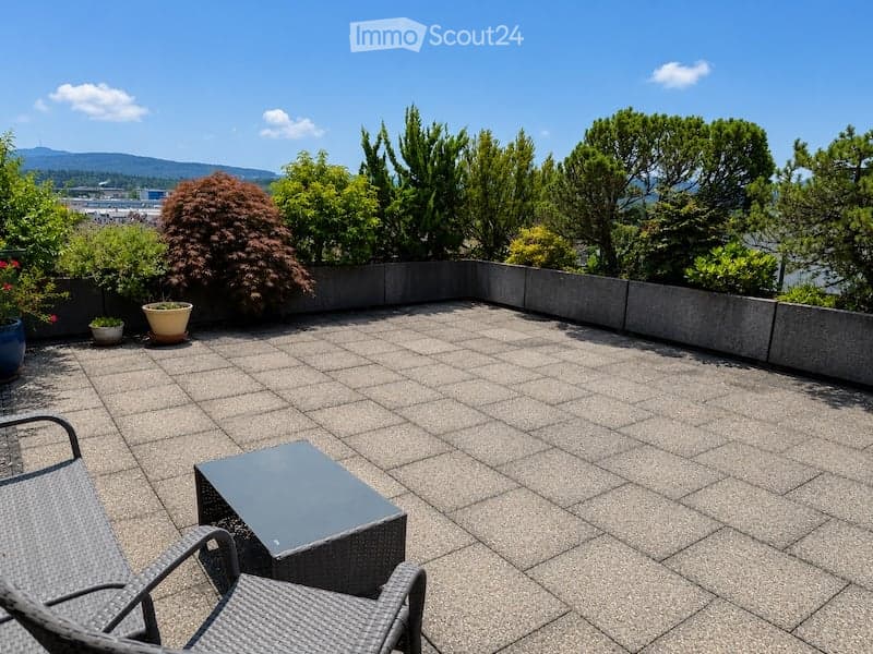 Spacious balcony with tiled floor, low concrete walls, and scenic view of trees and mountains. Includes a small table and two wicker chairs.