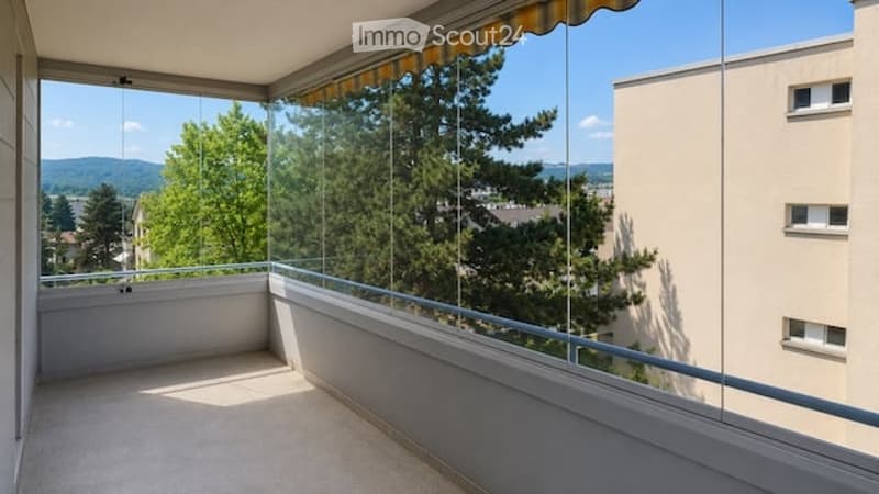 Balcony with glass railing, striped awning, overlooking green trees and distant hills.
