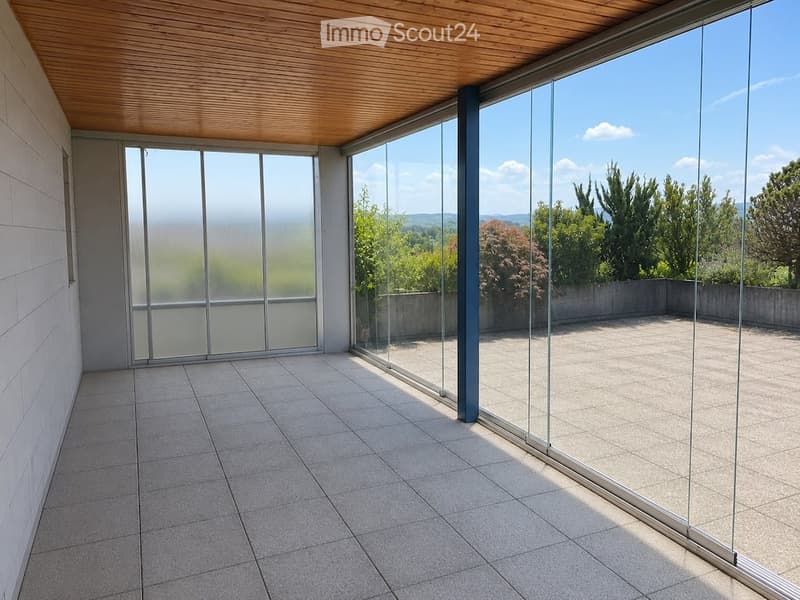 Modern balcony with large glass walls, tiled floor, wooden ceiling, and scenic mountain view.