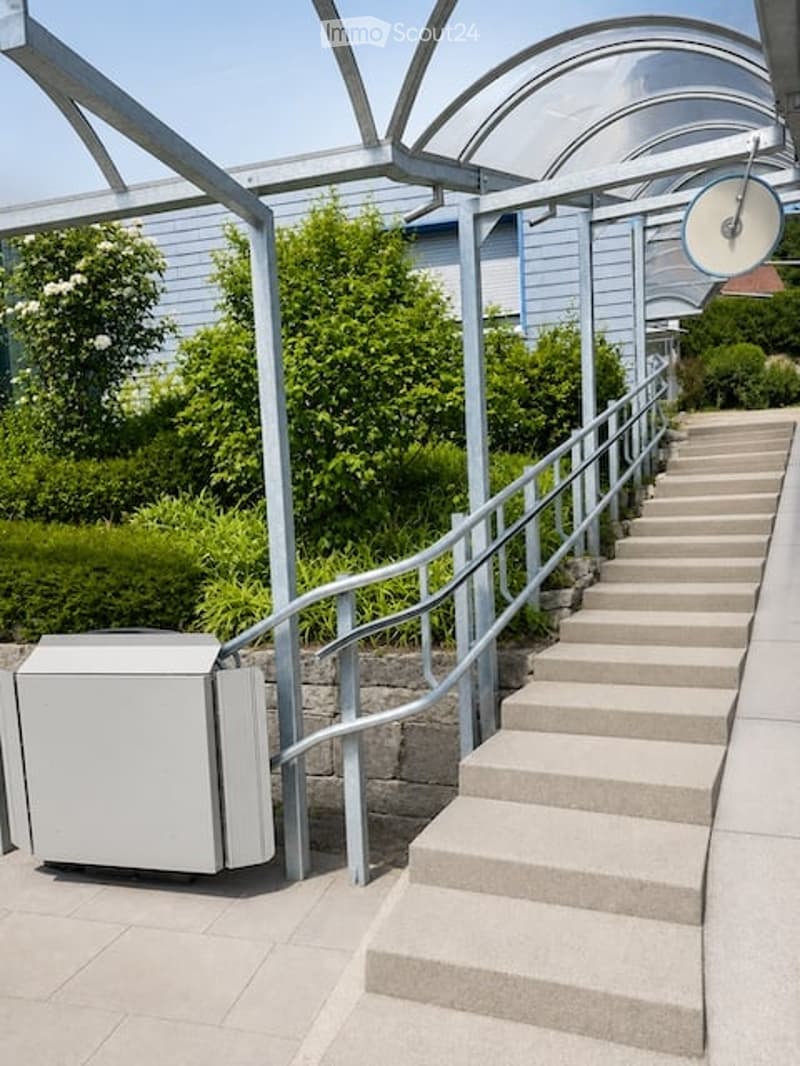 A modern outdoor staircase with metal railings and concrete steps, leading up to a covered walkway with a glass roof. The area is surrounded by greenery and features a small utility box on the left side.