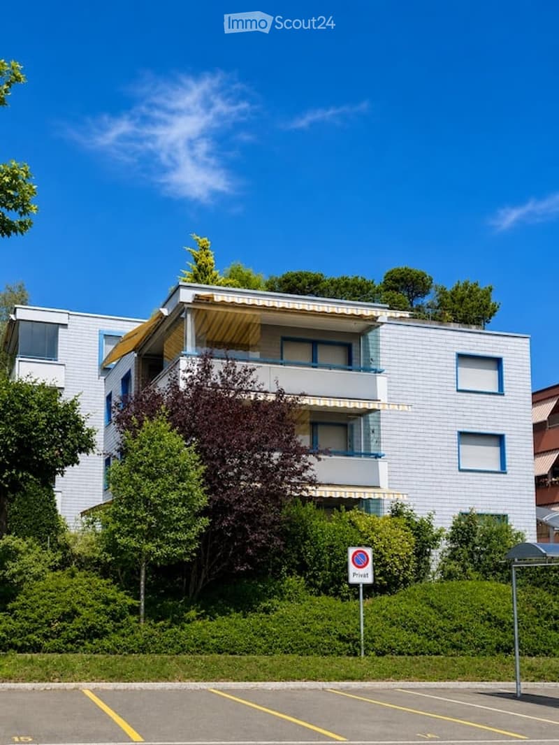 Modern multi-story apartment building with balconies, surrounded by greenery and a parking area.