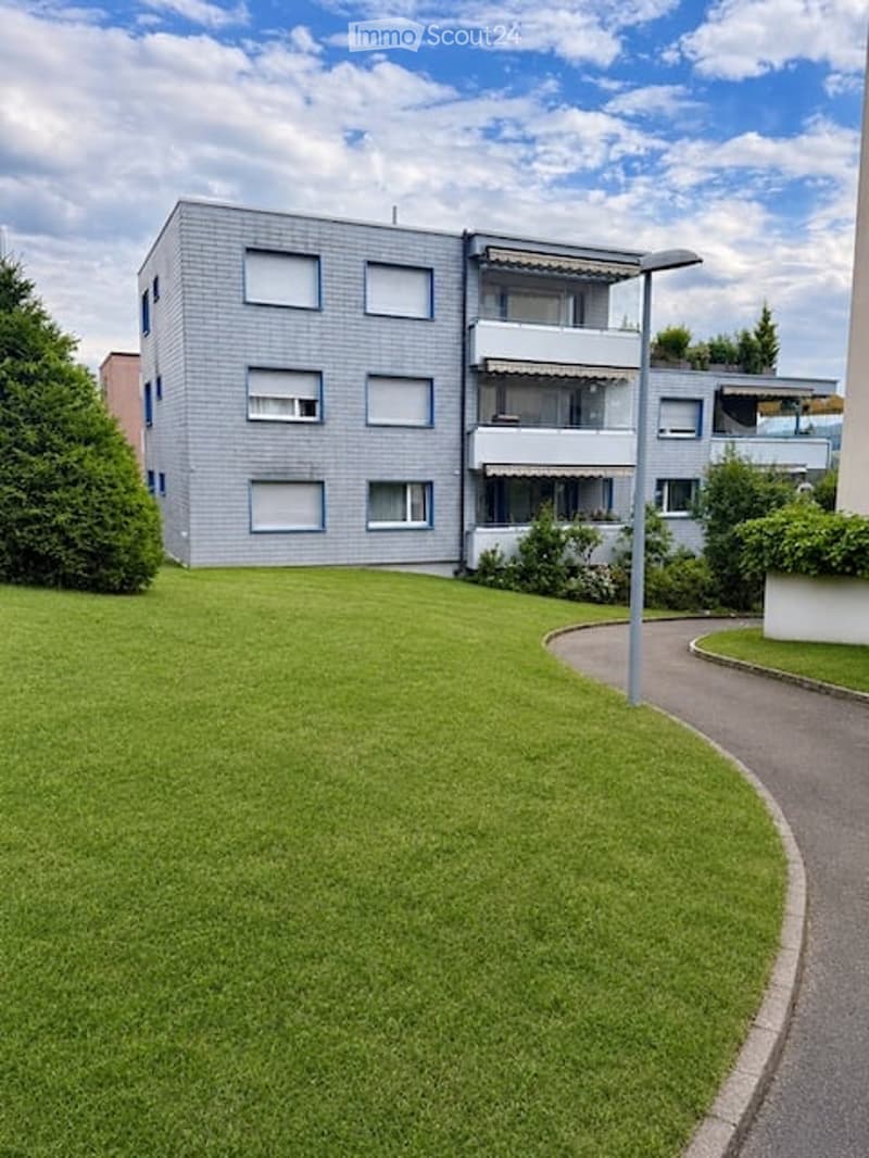 Modern multi-story apartment building with grey facade, multiple windows, and balconies. Surrounded by a well-maintained lawn and trees.