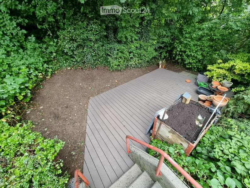 Wooden deck with a small raised garden bed, surrounded by greenery and plants. Includes gardening tools and pots.