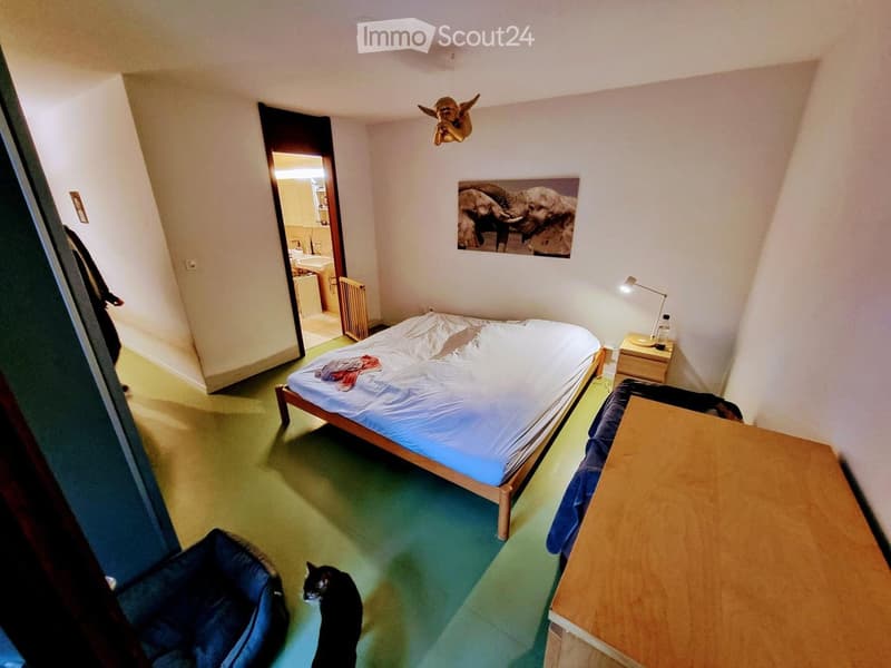 A small bedroom with a single bed, nightstand, wooden table, and a cat sitting on the floor. The room features a large mirror, a picture of elephants on the wall, and a decorative angel figurine hanging from the ceiling.