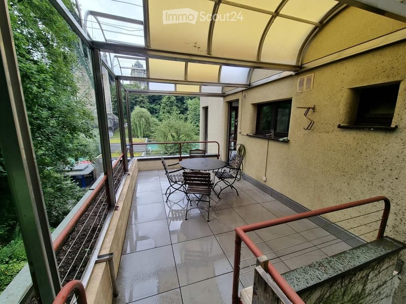Balcony with a table and chairs, surrounded by glass walls and a view of greenery.
