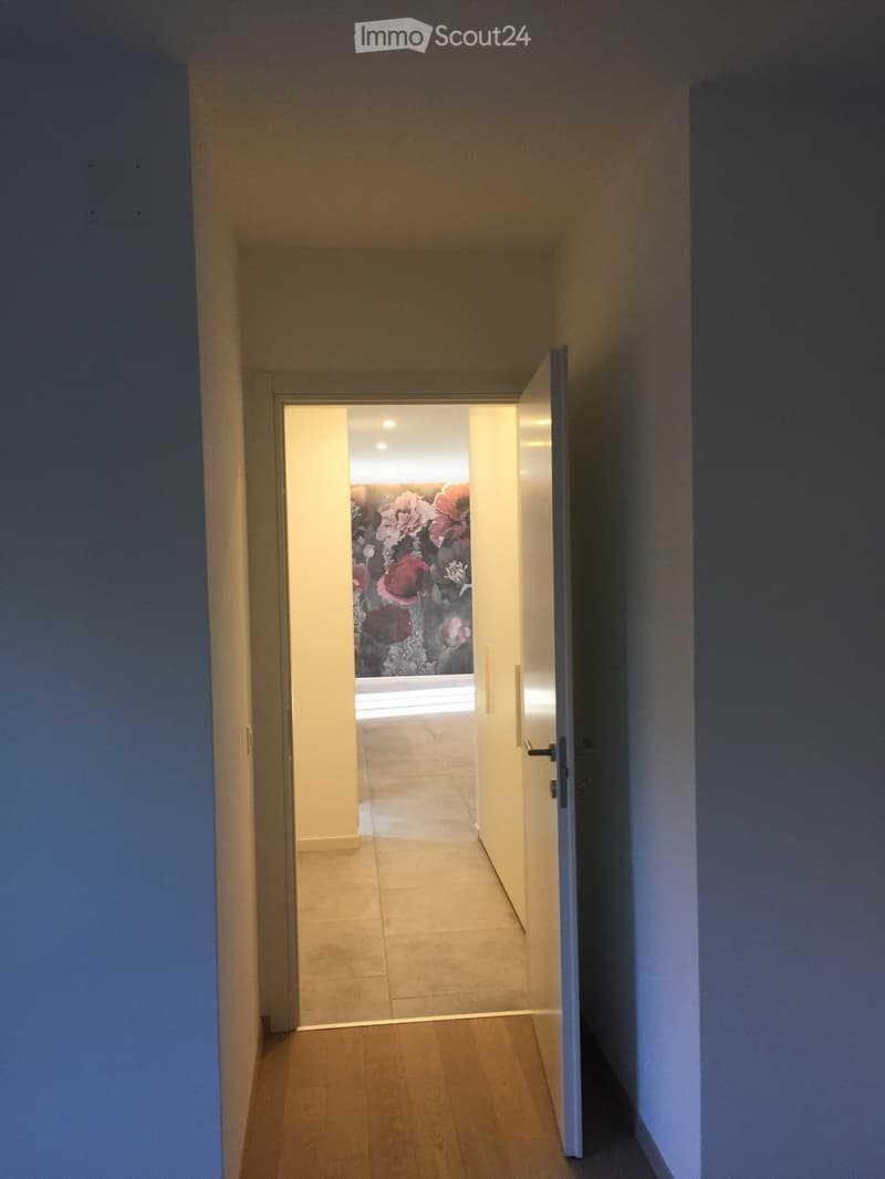 A narrow hallway with wooden flooring, light-colored walls, and an open door leading to another room with a floral wall design and tiled flooring.