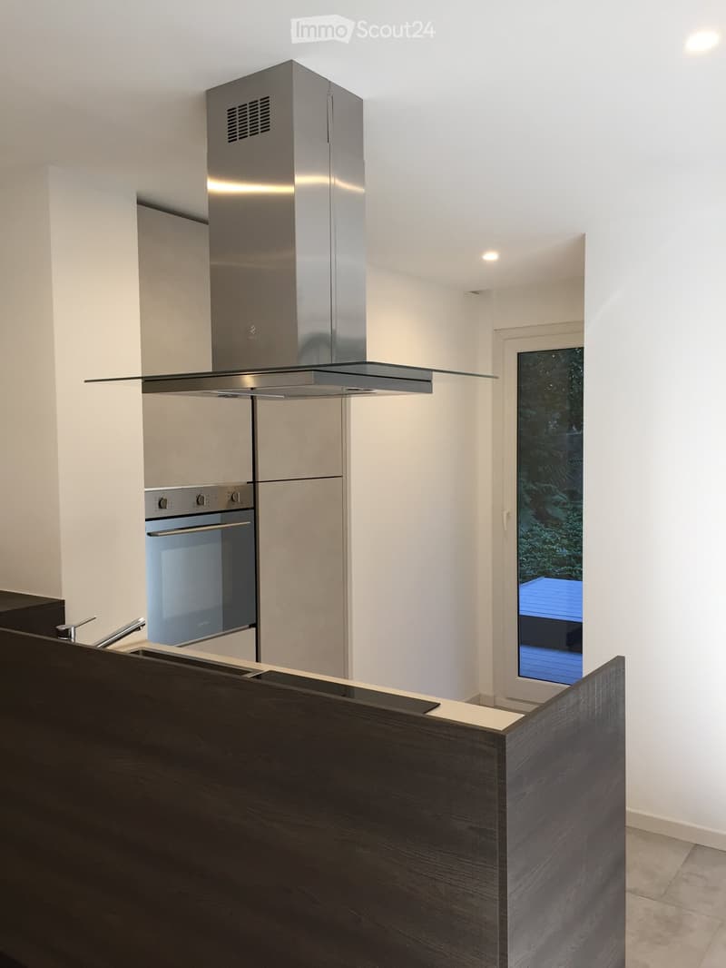 Modern kitchen with white walls, dark wooden countertop, stainless steel oven, and range hood. A large window offers a view of the outdoor area.