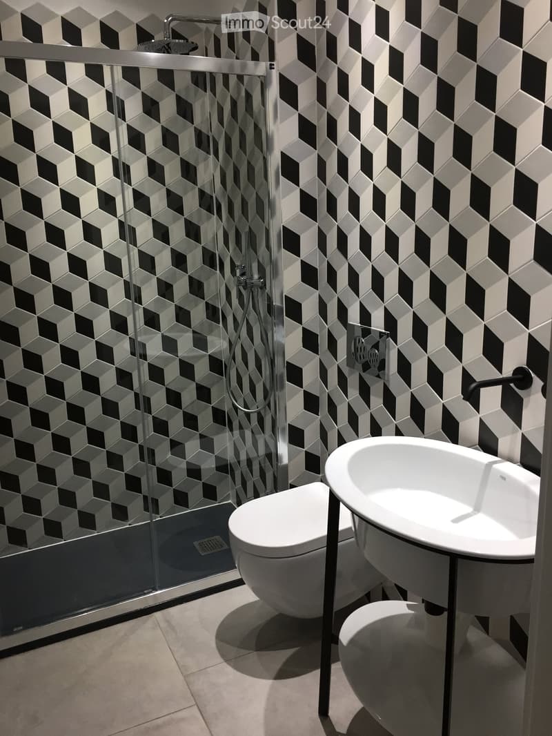 Modern bathroom with black and white geometric tile walls, a glass-enclosed shower, a toilet, and a sink with a sleek black faucet.