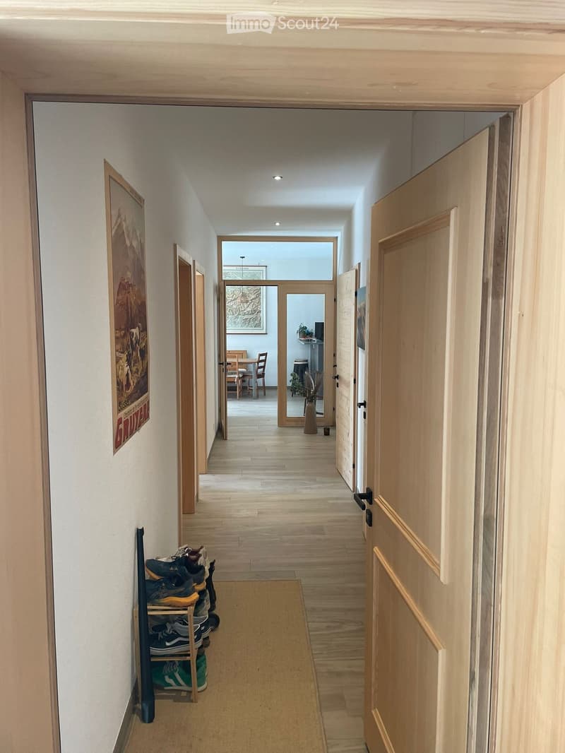 A long, well-lit hallway with wooden flooring, white walls, and several doors. A shoe rack with multiple pairs of shoes is visible on the left side, and a small rug lies in the center of the hallway. The hallway leads to an open area with a dining table a