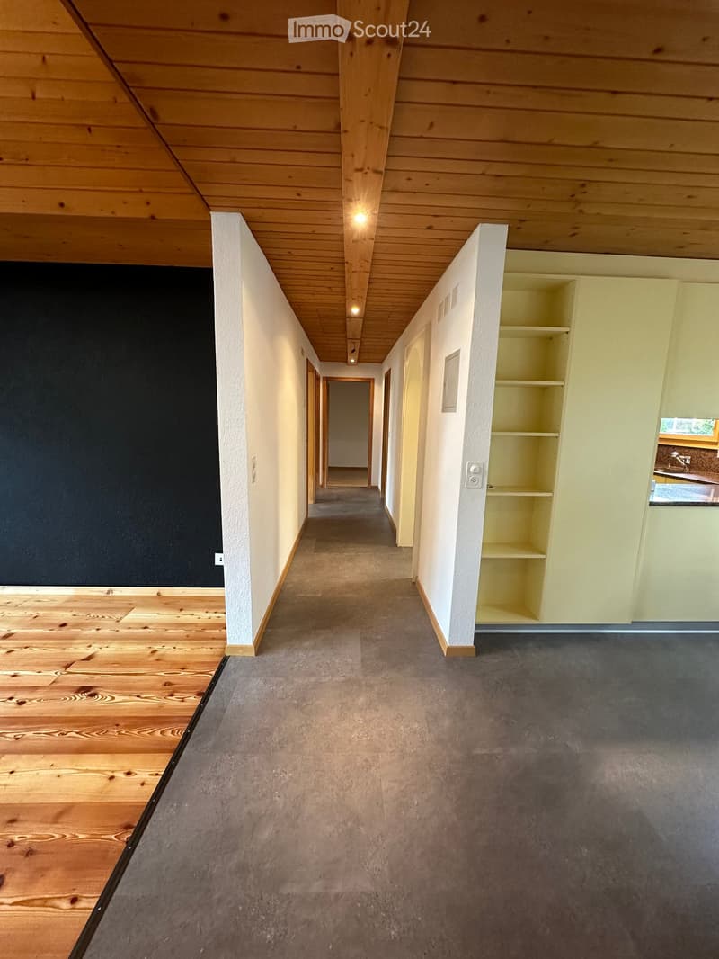Empty hallway with wooden flooring on one side and carpet on the other, white walls, and recessed lighting.