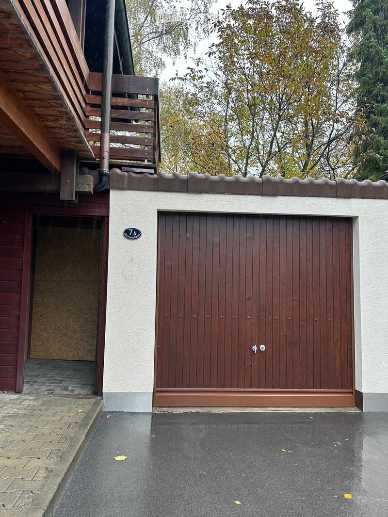 A modern house exterior with a large wooden garage door, a covered entrance, and a wooden balcony above. The facade is light-colored with a decorative wooden trim along the roofline.