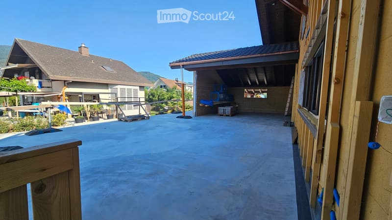 Outdoor patio area with concrete floor, wooden railing, and a covered garage area with storage items visible in the background.