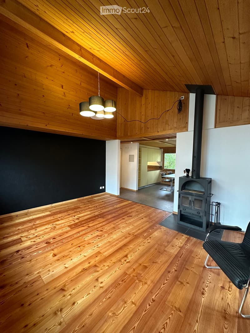 Spacious room with wooden floors, slanted wooden ceiling, pendant lights, and a fireplace. Adjacent kitchen area visible through an open doorway.