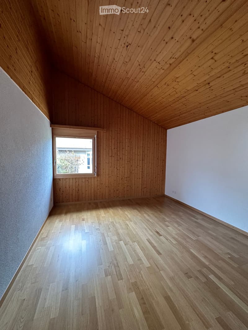 Empty room with wooden flooring and walls, featuring a slanted wooden ceiling and a single window with a view of the outside.