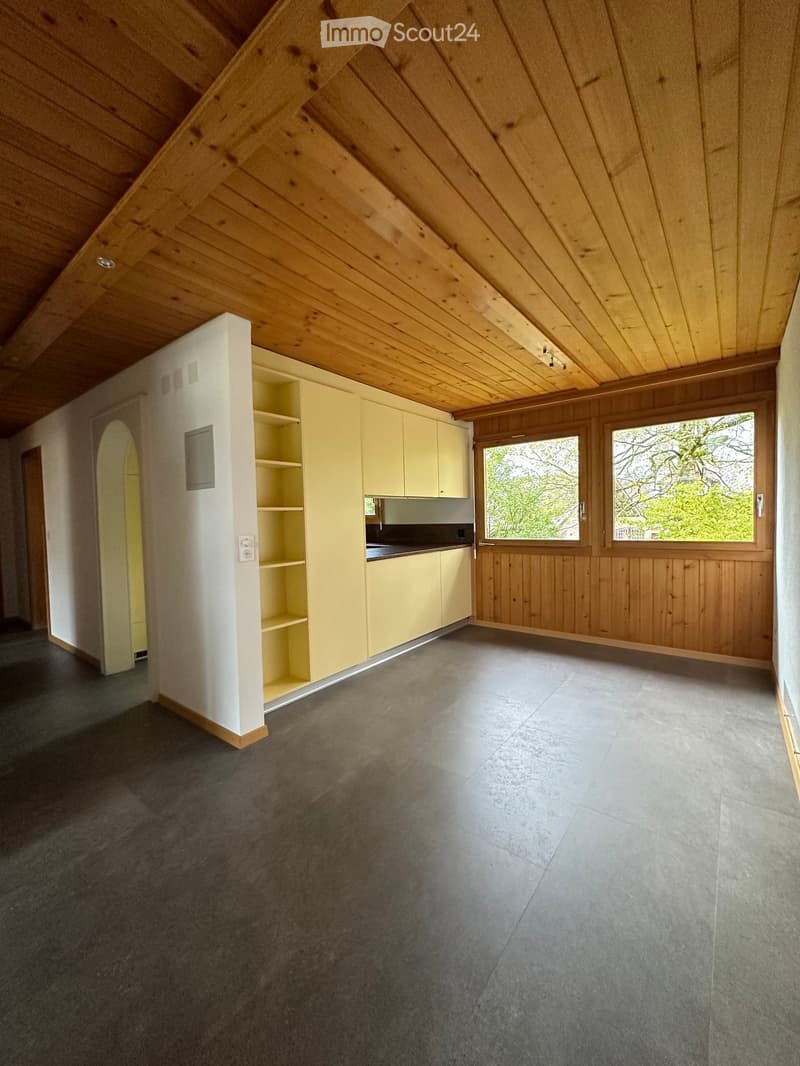 Empty attic space with wooden ceiling beams, light-colored cabinets, and large windows offering a view of greenery.