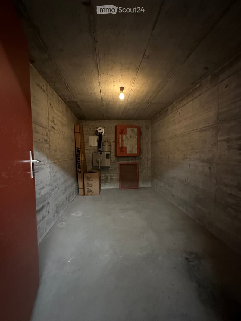 Empty concrete corridor with a single light fixture, a door on the left, and a few boxes and a fire extinguisher on the wall.