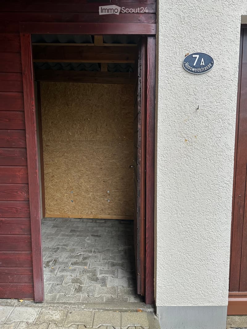 An open doorway leading into a wooden-framed interior space with a concrete floor, showing a partially visible interior with wooden paneling and a sign reading "7A Rossweidstrasse" on the adjacent wall.
