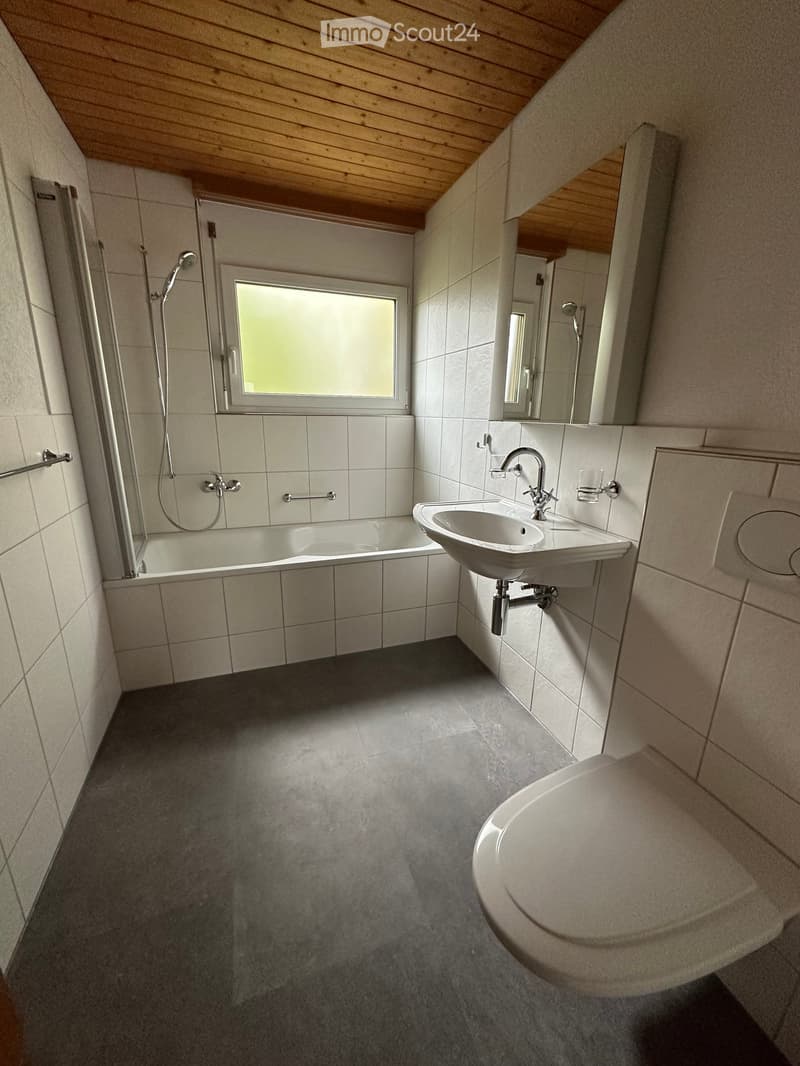 Modern bathroom with a bathtub, sink, toilet, and shower area. Features a large window, wooden ceiling, and tiled walls.