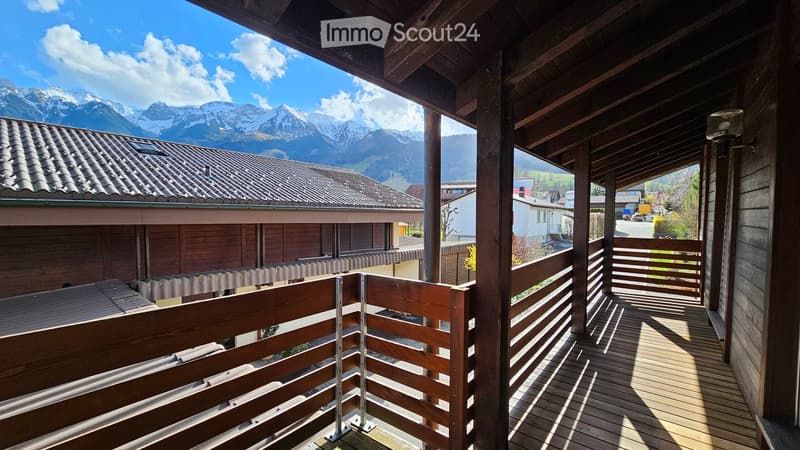 Wooden balcony with railing overlooking a scenic mountain view, with neighboring houses and a clear blue sky.