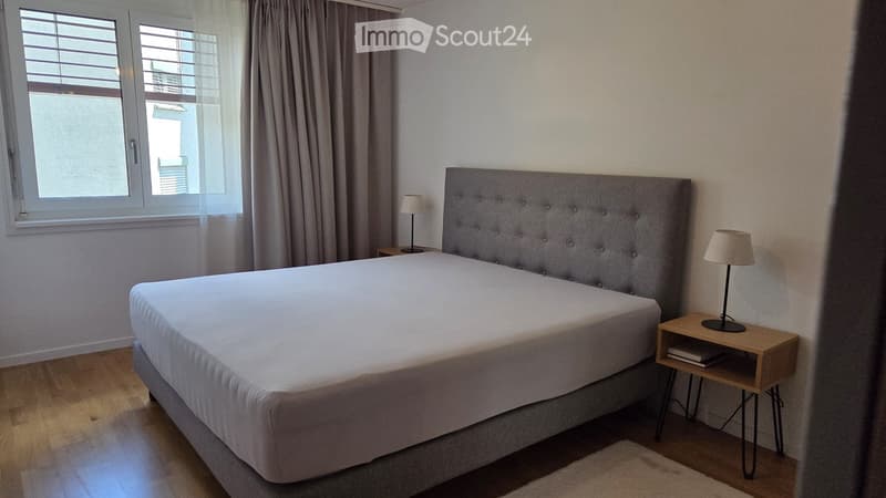 Modern bedroom with a large bed, grey headboard, wooden nightstand, table lamp, and sheer curtains on the window.