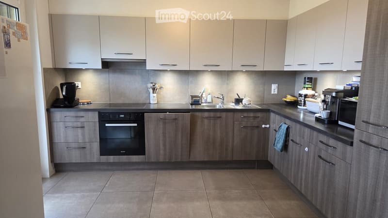 Modern kitchen with light grey cabinets, stainless steel appliances, and a tiled floor. Features an oven, microwave, coffee maker, and various kitchen items on the counter.