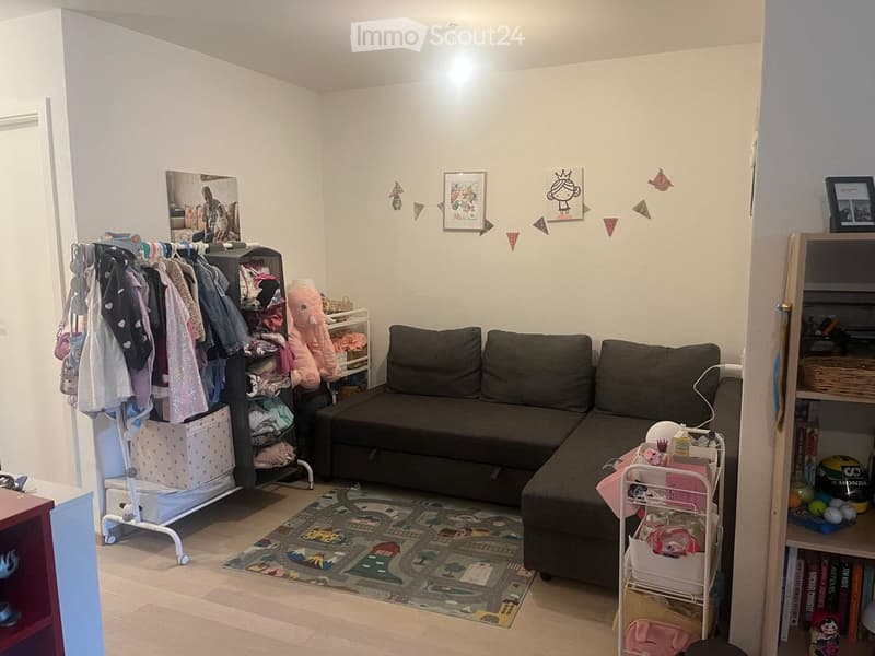 A cozy living room with a dark grey sectional sofa, a colorful play mat, a clothing rack, shelves with toys and storage bins, and a bookshelf with books and decorative items.