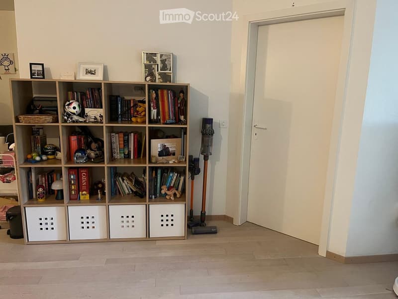 A small, organized room with a wooden shelf filled with books, toys, and framed pictures. A vacuum cleaner is placed near the door, and the floor is light-colored wood.