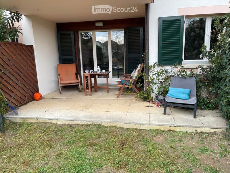 Outdoor patio area with a small table, four chairs, and a wooden fence. The patio is adjacent to a house with large glass doors and green shutters.