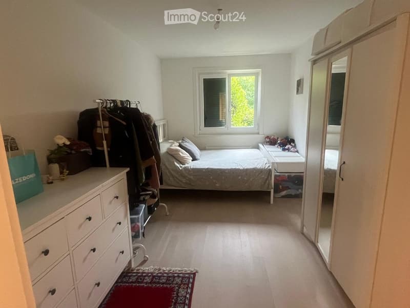 A small, well-lit bedroom with a single bed, nightstand, wardrobe, dresser, and a clothing rack. The room features white walls, a window with blinds, and a small rug on the floor.