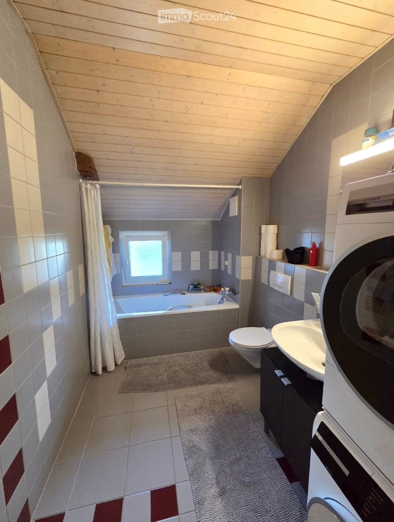 Modern bathroom with a bathtub, toilet, sink, and cabinet. Features a shower curtain, a window, and a light fixture above the sink. The floor has a tiled design with a small rug.