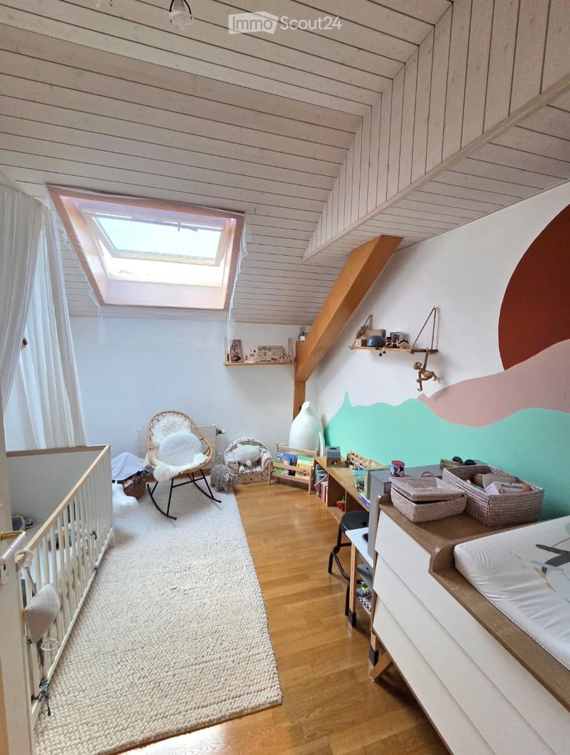 Cozy children's bedroom with wooden floors, slanted ceiling, skylight window, and colorful wall design. Features a baby crib, wicker chair, rocking chair, small armchair, bookshelf, table, storage baskets, and a bed.