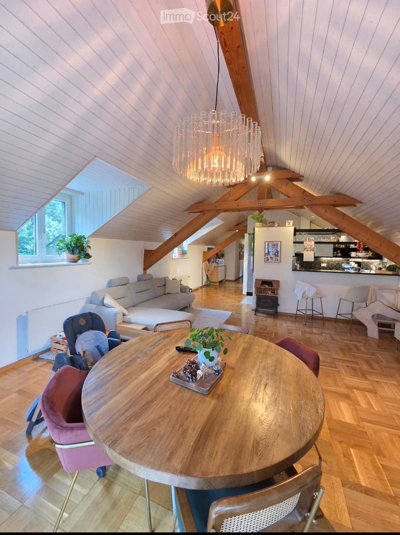 Open-plan living and dining area with wooden floors, exposed wooden beams, and a modern chandelier. Features a grey sofa, round wooden dining table with chairs, baby chair, and a small kitchen area with a counter and stools.