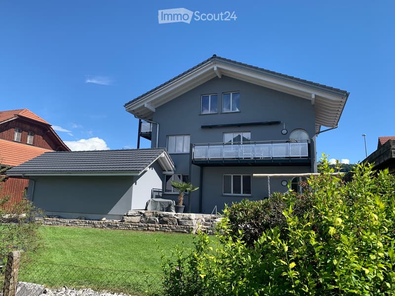 Two-story modern house with grey exterior, white trim, multiple windows, and a balcony on the upper level. The house features a well-maintained lawn and surrounding greenery.