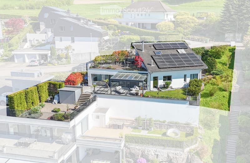 A modern, multi-level house with a combination of flat and sloped roofs, featuring large terraces, balconies, and solar panels on the roof. The property includes a well-maintained garden, outdoor seating areas, and a swimming pool.