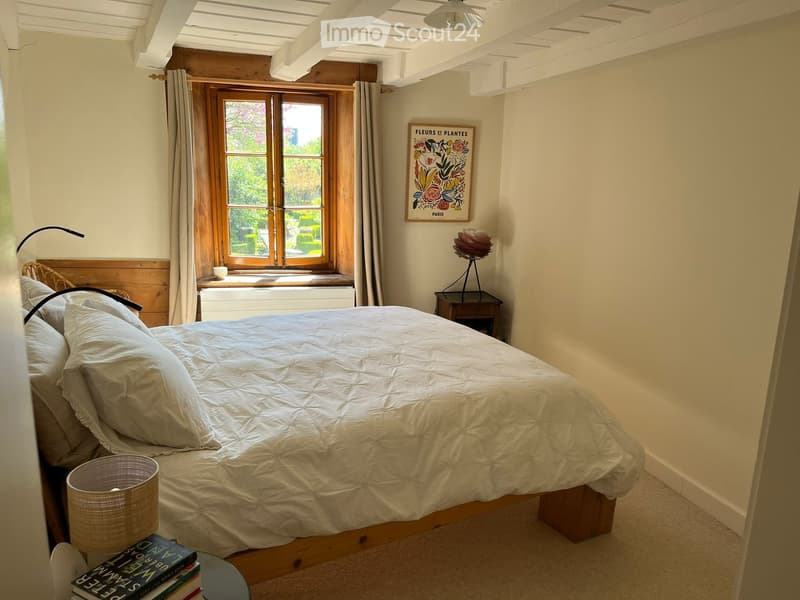 Bedroom. Cozy bedroom with a large bed, nightstand, lamp, books, and a decorative picture frame on the wall. The room features wooden beams and a large window with curtains.