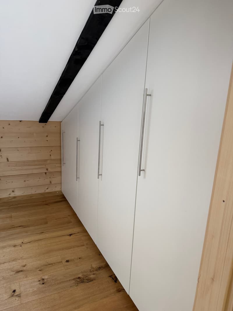 An empty attic space with wooden flooring and white walls, featuring built-in white cabinets with silver handles and a slanted ceiling with exposed beams.