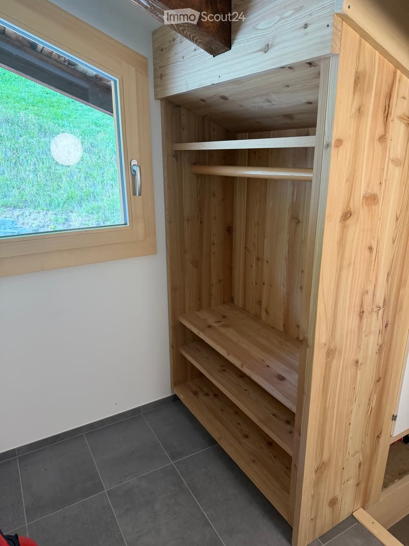 Empty room with wooden built-in shelving unit, dark tiled floor, and a window with a view of greenery outside.