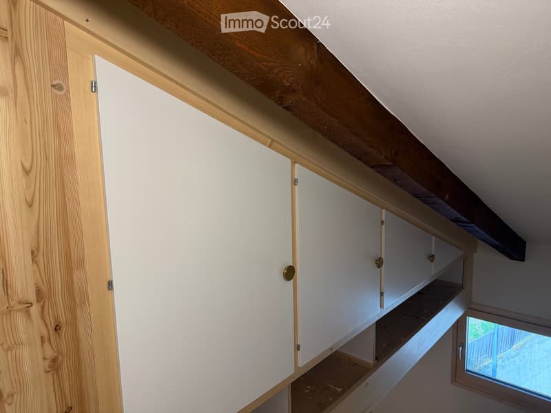 An empty attic space with exposed wooden beams, white built-in cabinets, and a small window showing a view of the outdoors.
