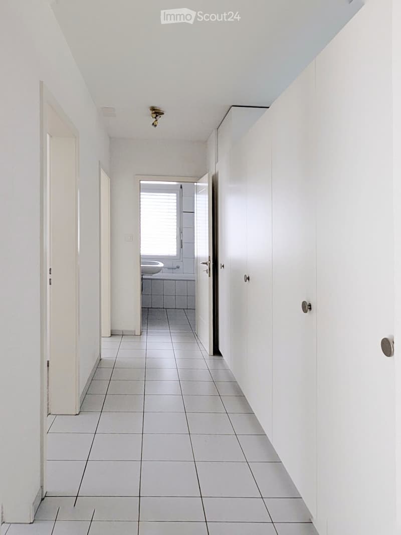 A clean, white-walled hallway with tiled flooring, leading to a bathroom visible at the end. The hallway features a recessed ceiling light and several doors along the walls.