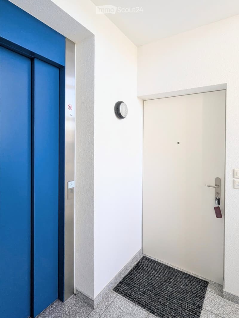 Modern hallway with white walls, tiled floor, blue elevator door, and a white apartment door with a keycard reader.