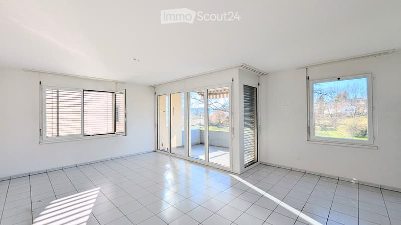 Empty room with white tiled flooring, white walls, and large windows with blinds. Features a sliding glass door leading to a balcony or outdoor area.